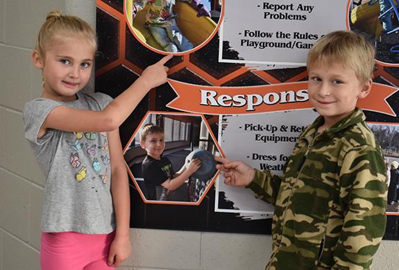 Riverview Elementary First Grade Students Miah Bartz and Keaton Kropp point to their photographs that were used for the new Playground Banner in the school’s atrium. This banner shows what students should be doing in order to be kind, safe, and responsible during recess.